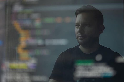 A man is looking at a computer screen displaying colorful lines of code. His reflection is visible on the screen, creating an overlay effect. The lighting is dim, and he appears focused and contemplative. The background is blurred, emphasizing the code and his reflection.