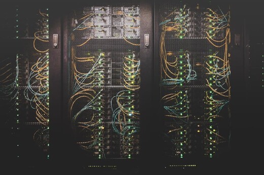 A dimly lit server room housing several black server racks, each filled with numerous computer servers. Colorful cables, mainly orange and yellow, are neatly plugged in, emitting small green and orange indicator lights, suggesting active connections and data flow.